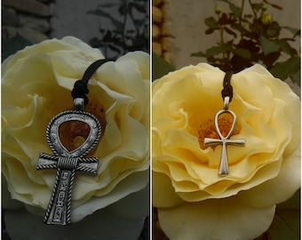 Pode incluir: Dois pingentes de Ankh prateados em cordões pretos, cada um descansando sobre uma rosa amarela. O Ankh à esquerda tem detalhes intrincados e uma superfície texturizada. O Ankh à direita é um design mais simples.