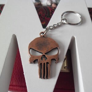 Pode incluir: Um chaveiro de caveira de cor cobre com uma corrente e um anel prateados. A caveira tem um design detalhado com uma superf&iacute;cie texturizada em relevo.