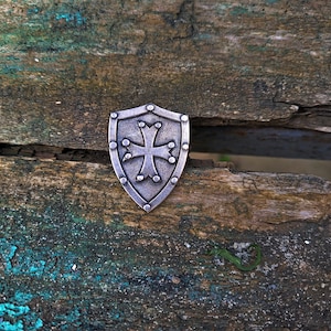 Puede incluir: Un escudo plateado con un diseño de cruz sobre una superficie de madera desgastada. El escudo tiene una cruz en relieve y está rodeado de pequeños remaches.