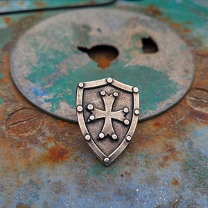 May include: A silver shield with a cross in the center, on a rusty metal surface.