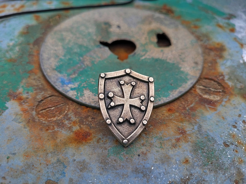 May include: A silver shield with a cross in the center, on a rusty metal surface.