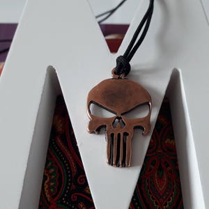 Pode incluir: Um pingente de caveira de cor cobre com um cord&atilde;o preto. A caveira tem um design detalhado com uma mand&iacute;bula proeminente e uma superf&iacute;cie texturizada.
