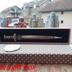 Puede incluir: Una espada decorativa en una caja de regalo de papel. La espada tiene una empu&ntilde;adura y una hoja de color bronce. La caja es marr&oacute;n con una tapa estampada en gris y blanco. Modelos de iglesias en miniatura en el fondo.