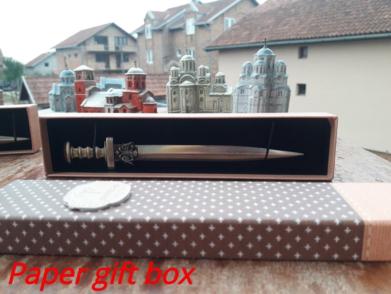 Puede incluir: Una espada decorativa en una caja de regalo de papel. La espada tiene una empu&ntilde;adura y una hoja de color bronce. La caja es marr&oacute;n con una tapa estampada en gris y blanco. Modelos de iglesias en miniatura en el fondo.