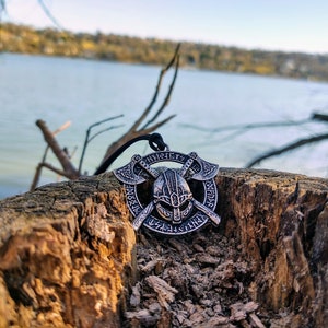 May include: Silver-colored Viking pendant featuring a helmet, crossed axes, and runic inscriptions. The pendant hangs on a black cord. The image is set against a natural backdrop of a tree stump and a body of water.