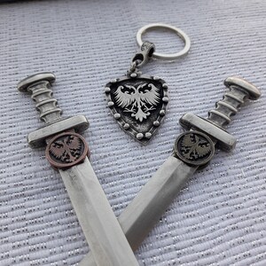 May include: Two silver swords with eagle emblems on the handles, crossed over each other. A silver shield with an eagle emblem hangs from a silver key chain.