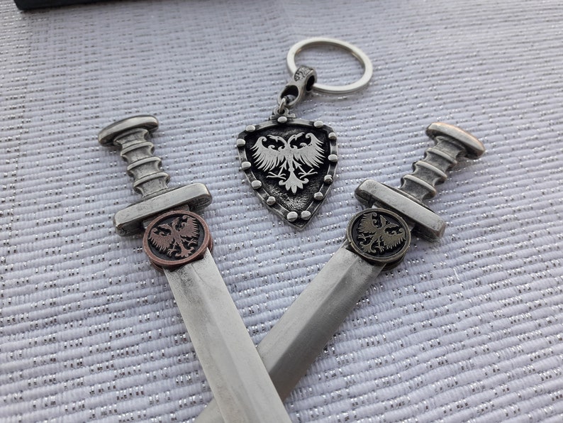 May include: Two silver swords with eagle emblems on the handles, crossed over each other. A silver shield with an eagle emblem hangs from a silver key chain.