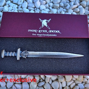 Puede incluir: Un abrecartas con forma de espada plateada en una caja burdeos. El abrecartas tiene un mango detallado y una hoja puntiaguda. La caja est&aacute; forrada con espuma negra y tiene la inscripci&oacute;n "SWORD LETTER OPENER" en la tapa.