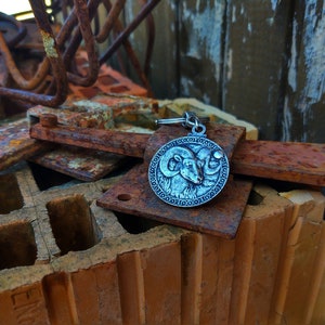 May include: A silver-toned keyring with a circular charm featuring a detailed embossed image of a ram's head. The keyring is resting on a pile of rusty metal and brick.