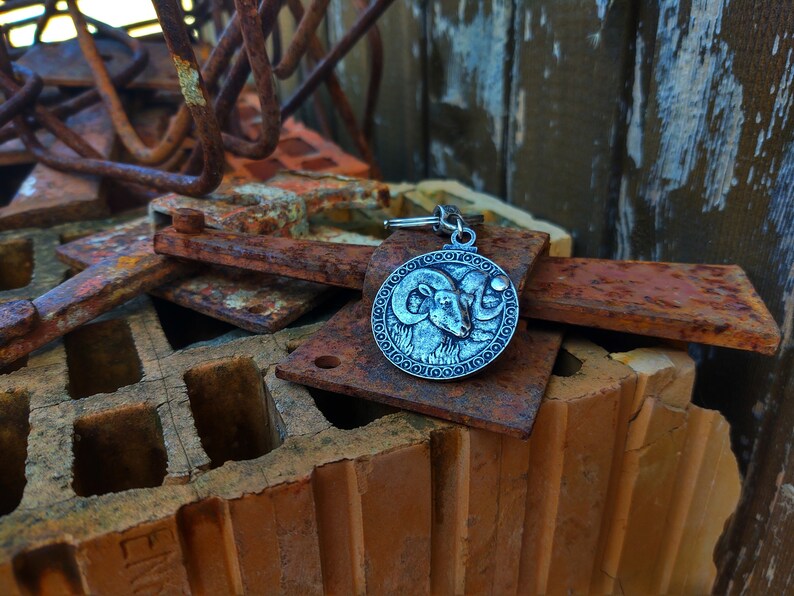 May include: A silver-toned keyring with a circular charm featuring a detailed embossed image of a ram's head. The keyring is resting on a pile of rusty metal and brick.