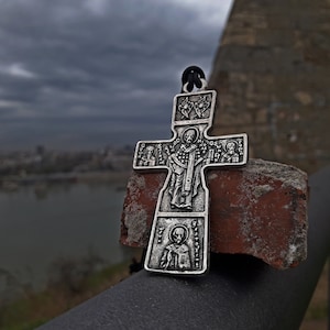 May include: A silver cross pendant with a detailed design featuring multiple figures. The cross is resting on a weathered brick against a backdrop of a cloudy sky and a river.