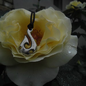 Puede incluir: Un colgante de plata con forma de gancho cuelga de un cordón negro. El colgante descansa sobre una rosa blanca con gotas de agua.