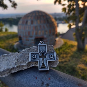May include: A silver cross pendant with an ornate design, resting on a grey rock. The cross is in focus, while the background is blurred, showing a brick dome structure and green foliage.