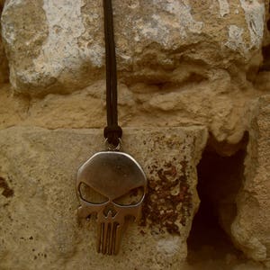 Pode incluir: Um colar com um pingente de caveira de prata com um cord&atilde;o preto pendurado em uma parede de pedra &aacute;spera.