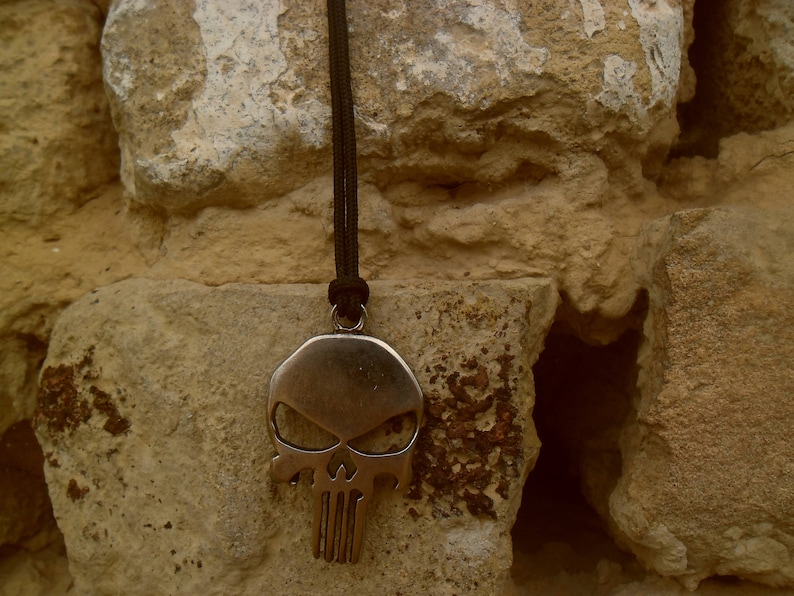 Pode incluir: Um colar com um pingente de caveira de prata com um cord&atilde;o preto pendurado em uma parede de pedra &aacute;spera.