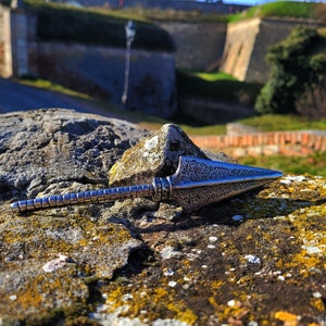 May include: A silver-colored arrowhead pendant with a textured surface. The pendant is lying on a rock with moss and lichen.