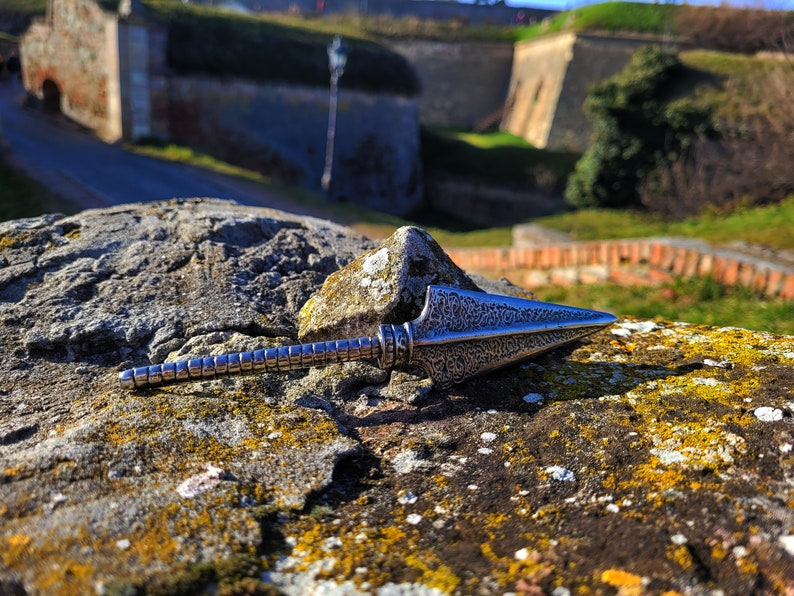 May include: A silver-colored arrowhead pendant with a textured surface. The pendant is lying on a rock with moss and lichen.
