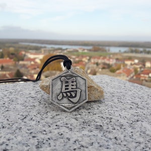 May include: A silver hexagonal pendant necklace with a black Chinese character on it. The necklace is on a stone surface with a blurry background of a town.
