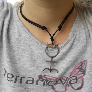 May include: A black cord necklace with a silver pendant featuring a stylized symbol with a circle, a cross, and three curved lines at the bottom.