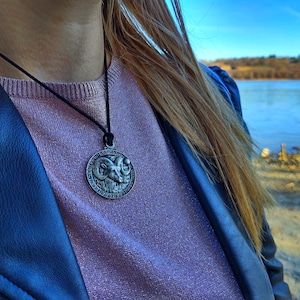 May include: A silver pendant necklace with a ram's head design. The pendant is round and has a black cord.