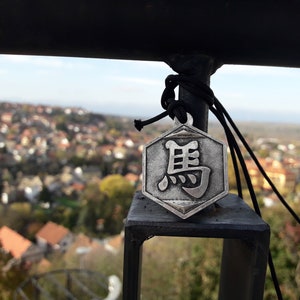 May include: A silver hexagonal pendant with a Chinese character on it. The pendant is hanging from a black cord and is tied to a black metal railing. The pendant is in focus, while the background is blurred.