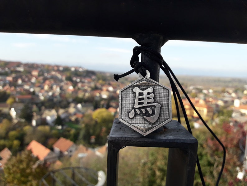 May include: A silver hexagonal pendant with a Chinese character on it. The pendant is hanging from a black cord and is tied to a black metal railing. The pendant is in focus, while the background is blurred.