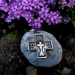 May include: A silver metal cross keyring with a circle design on each side of the cross. The keyring is resting on a grey rock with purple flowers in the background.