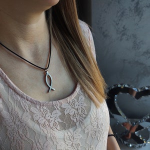Puede incluir: Un collar con un colgante de pescado plateado en un cordón negro. El collar lo lleva una persona que lleva una blusa de encaje rosa.