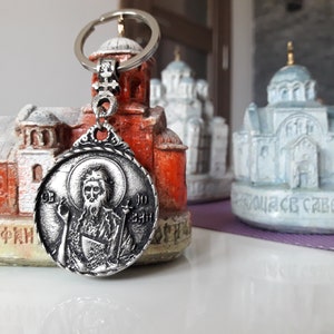 Könnte beinhalten: Silberner Schlüsselanhänger mit einem runden Symbol, das eine religiöse Figur zeigt. Das Symbol hat einen silbernen Heiligenschein und die Figur hält einen Stab. Der Schlüsselanhänger hängt an einem silbernen Ring.