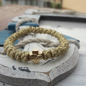 Peut inclure: Un bracelet tressé de couleur or avec un charm en forme de croix. Le bracelet est fait d'un matériau épais et texturé et comporte une petite croix détaillée. Le bracelet repose sur une ancre décorative.