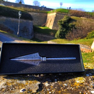 May include: A silver-colored arrowhead-shaped keychain in a black velvet box. The keychain is detailed with a textured pattern and is displayed on a rock with a blurred background of a stone wall and green grass.