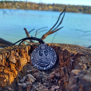 May include: A silver pendant necklace with a circular design featuring a stylised bird with outstretched wings. The pendant is on a black cord and is resting on a weathered tree stump.