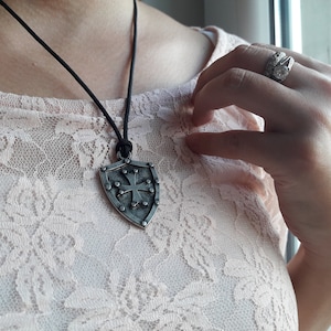 May include: A silver shield pendant with a cross design hangs from a black cord necklace. The pendant has a textured surface and is worn by a person.
