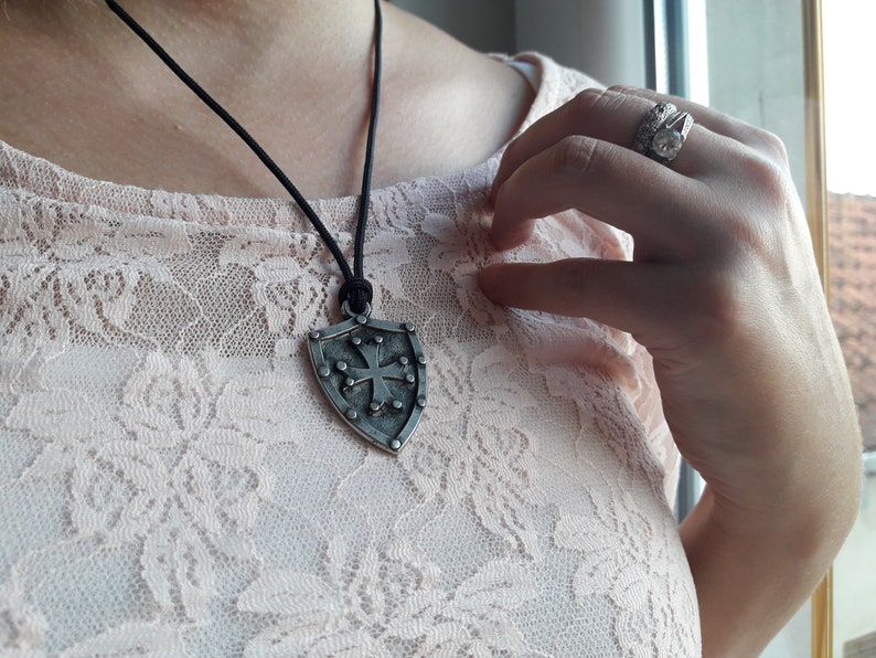 May include: A silver shield pendant with a cross design hangs from a black cord necklace. The pendant has a textured surface and is worn by a person.