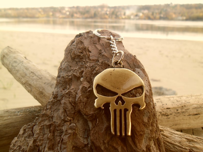 Pode incluir: Um chaveiro de metal dourado com um design de caveira estilizado. O chaveiro est&aacute; pendurado em uma corrente prateada e est&aacute; apoiado em um peda&ccedil;o de madeira &agrave; deriva.