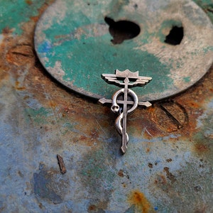 May include: A silver metal pendant with a cross, a snake, and wings. The pendant is on a rusty metal surface.