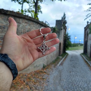 Puede incluir: Un llavero plateado con un nudo celta. El llavero presenta un dise&ntilde;o de nudo de cuatro puntas, suspendido de un anillo de metal. El fondo muestra un camino empedrado y muros de piedra.