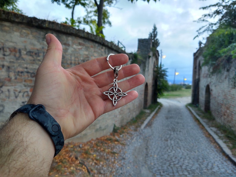 Puede incluir: Un llavero plateado con un nudo celta. El llavero presenta un dise&ntilde;o de nudo de cuatro puntas, suspendido de un anillo de metal. El fondo muestra un camino empedrado y muros de piedra.