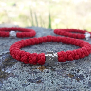 May include: Three red knotted bracelets with silver cross charms. The bracelets are on a gray, textured surface.