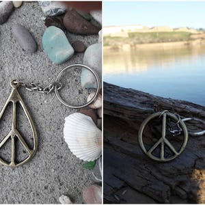 May include: A silver peace sign keychain with a silver ring and a gold peace sign charm. The keychain is on a gray background with small stones and a white seashell.