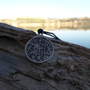 May include: A silver pendant with a circular design featuring a geometric pattern and symbols. The pendant is hanging from a black cord and is resting on a piece of weathered wood.