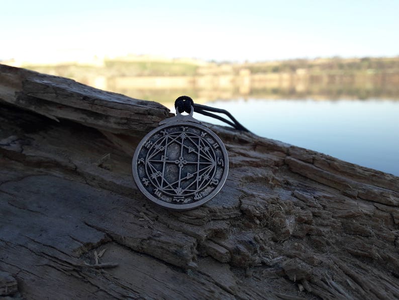 May include: A silver pendant with a circular design featuring a geometric pattern and symbols. The pendant is hanging from a black cord and is resting on a piece of weathered wood.