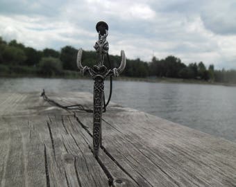 Könnte beinhalten: Eine silberne Schwertpendantkette mit filigranen Details und einer schwarzen Schnur. Der Anhänger liegt auf einer verwitterten Holzoberfläche in der Nähe eines Gewässers.