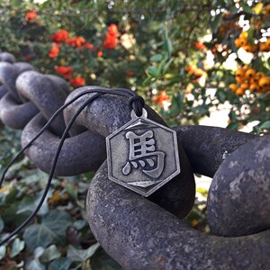May include: A silver hexagonal pendant with a Chinese character on a black cord, hanging from a rusty chain link fence. The pendant is in focus, while the background is blurred.