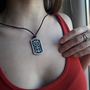 Op de afbeelding: Een zilveren ketting met een zwarte koord. De hanger is een rechthoekige tag met een symbool dat lijkt op een gestileerde vrouwelijke figuur.