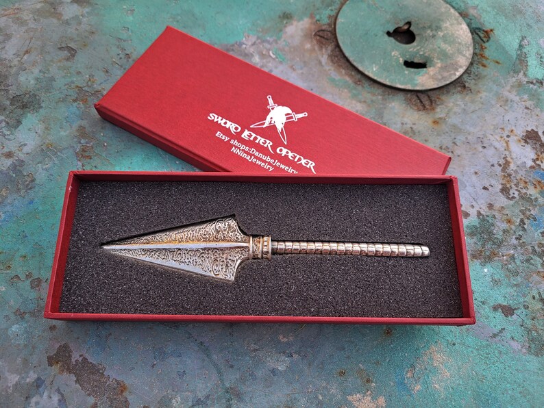 May include: A silver bottle opener in the shape of a spearhead, with a textured surface, in a red velvet box. The box has the text "SWORD LETTER OPENER" and "Etsy shop: DanubeJewelry" printed on the lid.