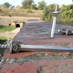 May include: Three miniature silver swords with ornate handles and a cross design on the pommel. The swords are lying on a brick wall.
