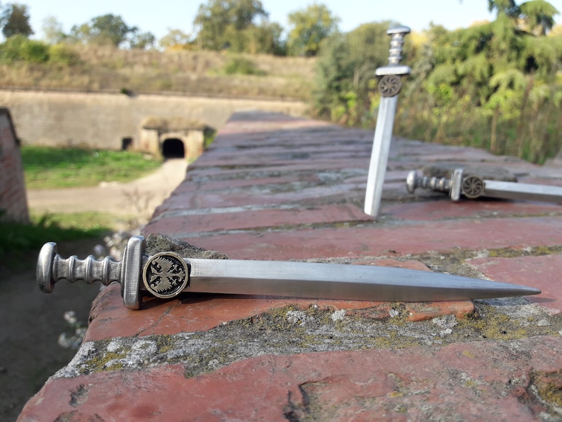 May include: Three miniature silver swords with ornate handles and a cross design on the pommel. The swords are lying on a brick wall.