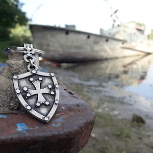 May include: A silver-toned keychain featuring a shield with a cross design. The keychain is resting on a rusty metal surface with a blurred background of a boat and water.