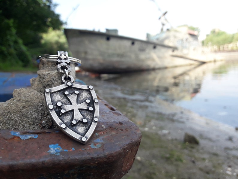 May include: A silver-toned keychain featuring a shield with a cross design. The keychain is resting on a rusty metal surface with a blurred background of a boat and water.